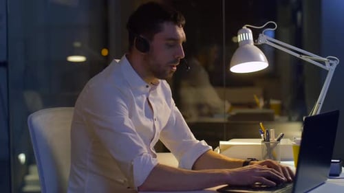 Young hispanic businessman typing on laptop with headset overworking late at night office