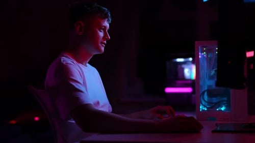 Fröhlicher junger Mann, der nachts Videospiele auf einem Desktop-Computer spielt, beleuchtet von Vibrant