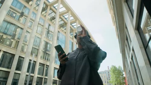 Black Woman Looking at Smartphone and Drinking Coffee, Walking Around Urban City