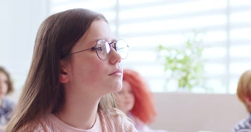 Close Up of Face of Cute Teen Clever Schoolgirl in Glasses Sitting in Classroom and Rising Hand to