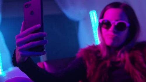 Beautiful Young Girl Taking Selfie with Her Phone on Bed with Neon Colors Room