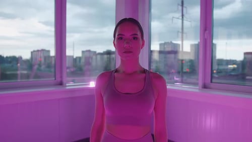Portrait of a Beautiful Young Girl in a Fitness Studio with Lilac Neon Lights and Large Windows