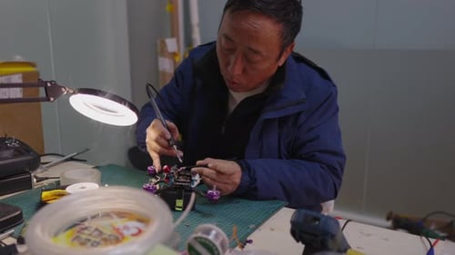 Asian Man Carefully Soldering Small Electronic Components Onto Circuit Board Under a Magnifying Lamp