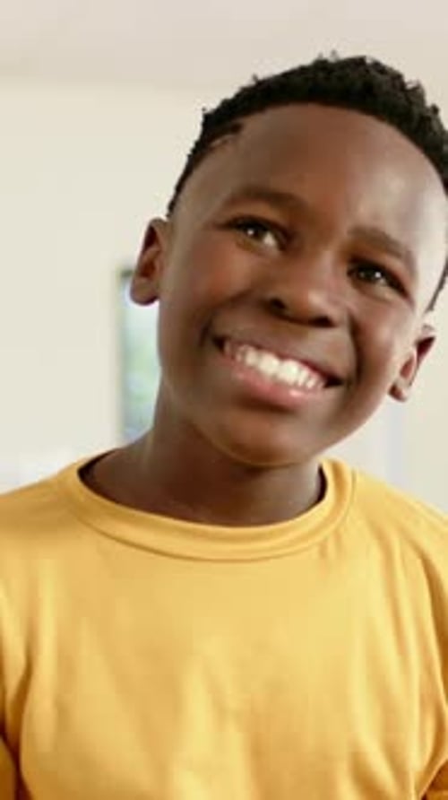 Vertical video: Smiling boy tilting head, shifting gaze at off-camera prompt at home in yellow tee