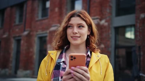 Redhead Girl in Yellow Raincoat Holding Mobile Phone and Smiling on Street in City Outdoor Portrait