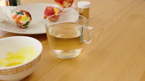 Hand places a colorful, cloth-wrapped Easter egg onto a plate, with bowls of yellow dye and a glass