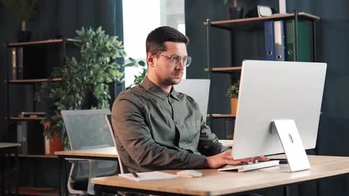 Handsome Man Working on Laptop Computer Sitting Behind Desk in Office Room
