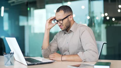 Young adult man suffering from headache while sitting at workplace at desk in modern office.