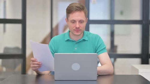 Man Reviews Documents at Desk with Laptop