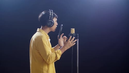 Side View Of Young Asian Boy With Headphone Singing Into A Condenser Microphone On Black Background