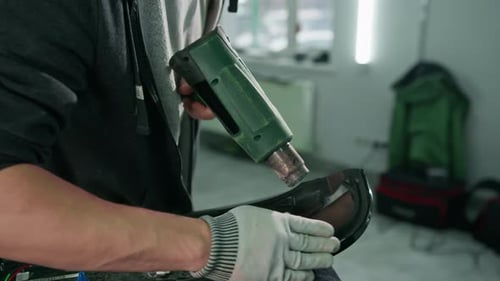 A worker covers a car spoiler with a film in the workshop using a hair dryer and a spatula close-up