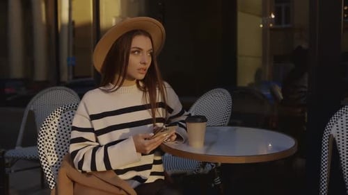 A Beautiful Young Girl in a Hat Sits in a Street Cafe and Uses a Mobile Phone to Scroll Through