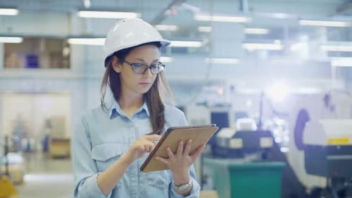 Una ingeniera industrial con un casco usa una tableta mientras camina por una gran fábrica.