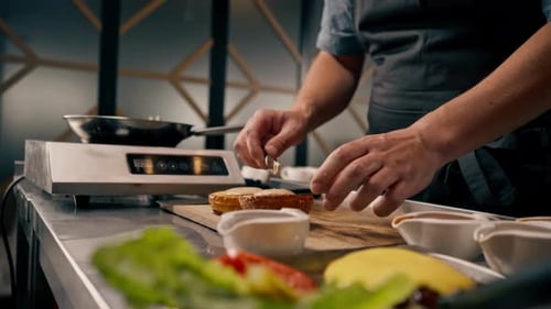 A close-up of a chef spreading sauce on a bun while cooking a burger in a professional kitchen
