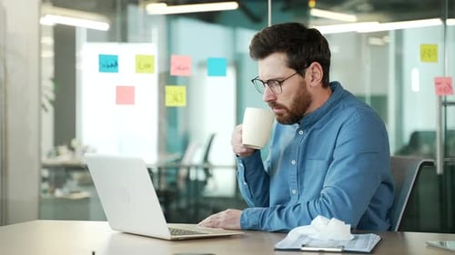 Sick businessman suffers from a cold while sitting at a desk at a workplace in a business office.