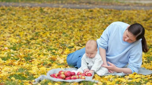 Mother Looks After Baby Girl Eating Red Apple on Park Lawn