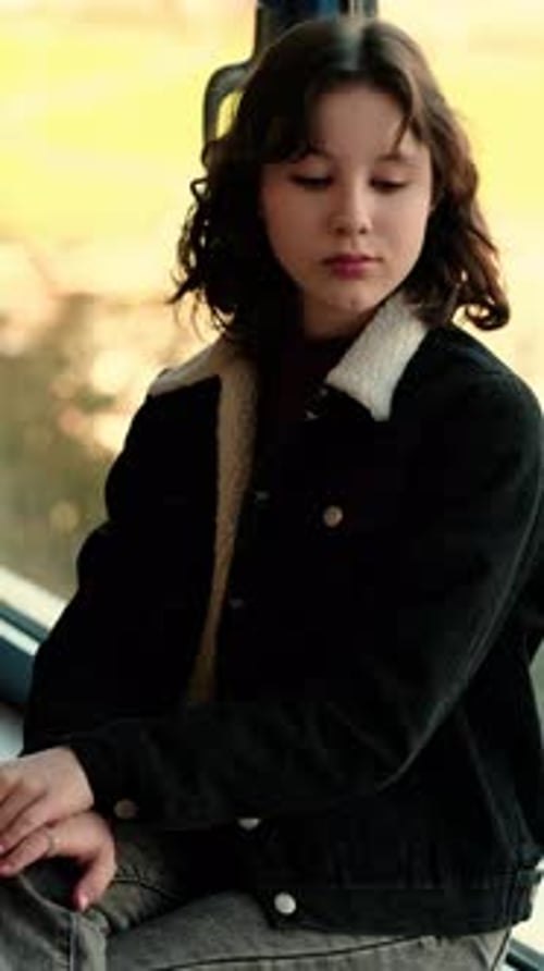 Teenage Girl in Warm Jacket Sits By the Window and Looks Down Sadly Thinking About Her Problems and