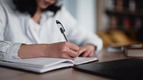 Woman Writes in Notebook Near Laptop at Desk