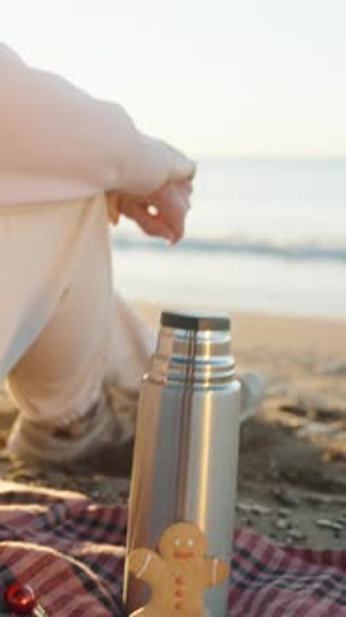 Vertical video. A young woman enjoys the Christmas morning sunrise by the sea, sitting on a plaid bl