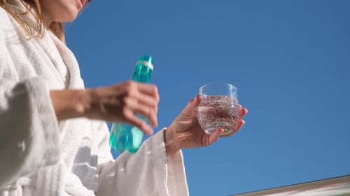 woman wearing bathrobe standing in the hotel balcony pouring mineral water into glass