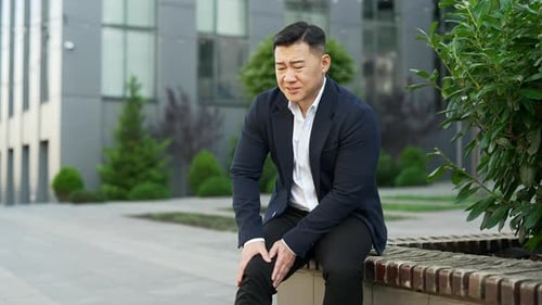 Asian man in formal suit suffers from painful severe knee joint pain sitting on a bench on street