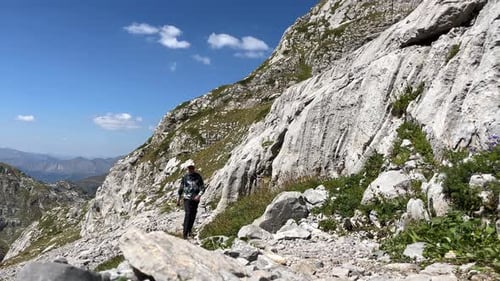 A Male Tourist with a Backpack Walks Along a Rocky Mountain Path on a Sunny Day Against the