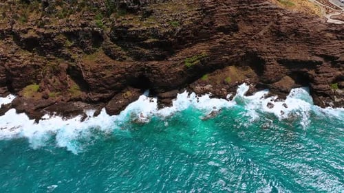 Dark bare rocks on the coast of the sea or ocean. Turquoise water hits the cliffs creating foam