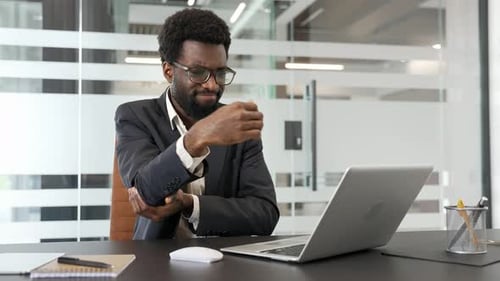 African american businessman in suit suffering from elbow pain sitting at desk in a business office.
