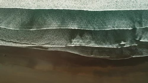 Aerial top view of ocean waves forming patterns on dark sandy shore