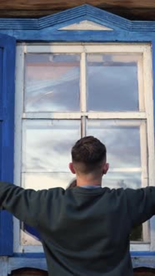 Vertikales Video: Ein Mann öffnet die Fensterläden an einem Fenster in einem alten Holzhaus