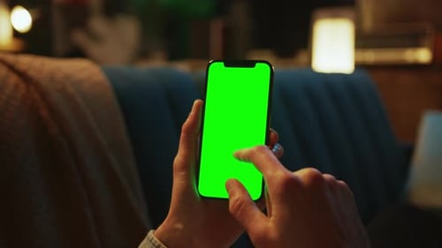 Close Up Hands Holding and Using Mobile Phone with a Vertical Green Screen Lying on a Sofa Indoors