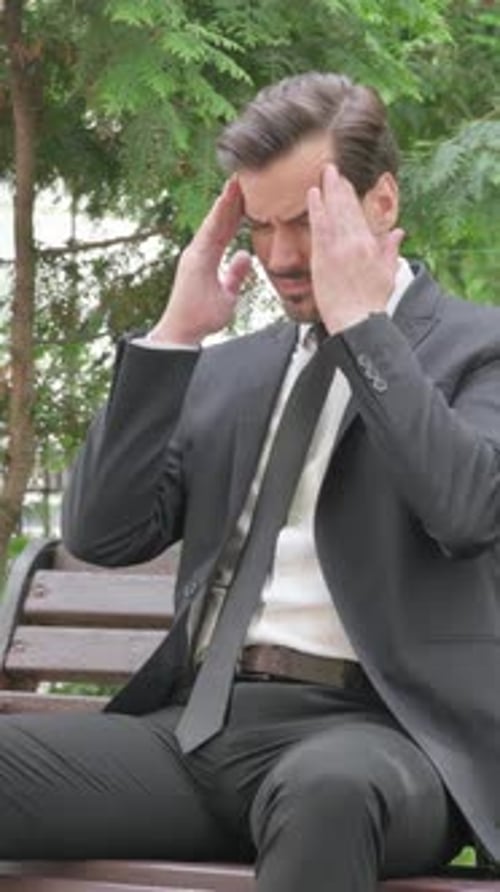 Stressed Man in Suit Massaging Temples on Park Bench