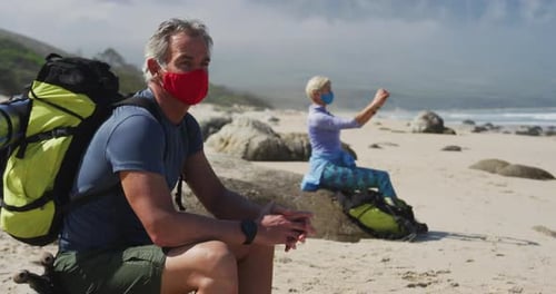 Senior hiker man wearing face mask with backpacks sitting on a rock and senior woman taking pictures