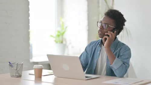 Young African Man Talking on Phone While Using Laptop in Office
