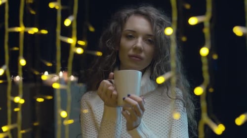 Woman Drinks from Mug by Window with Lights