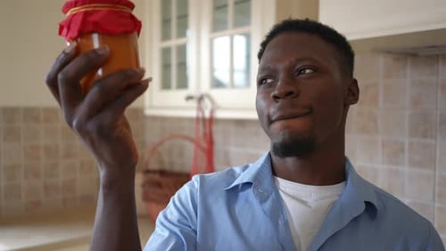Jeune homme souriant admirant la confiture d'orange en bocal debout dans la cuisine à l'intérieur