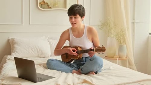 Woman Playing Ukulele on Bed with Laptop