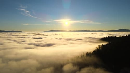 Aerial view of vibrant sunset over white dense clouds with dark mountain spruce forest trees.