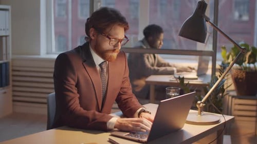 Young Male Office Worker Typing on Laptop at Office Desk in the Evening