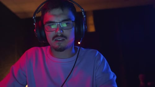 Man with headphones is playing videogames on laptop in room with neon lighting, front view. Male stu
