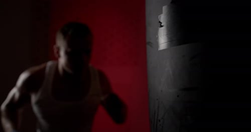 A man in a semi-dark hall, wearing hand wraps, hits a punching bag with quick and accurate punches.