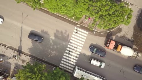 Vista aérea de cima para baixo da rua com carros em movimento e faixa de pedestres com pedestres cruzando.