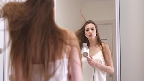Young Pretty Female Stands Near the Mirror and Dries Her Hair with a Hair Dryers, View of the