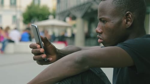 young african man types on smartphone sitting in the street