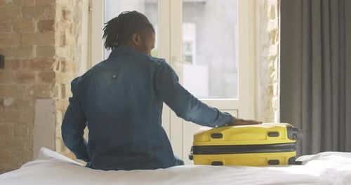 Back View of African American Young Man Sitting on Bed with Luggage Before Trip, Taking Yellow