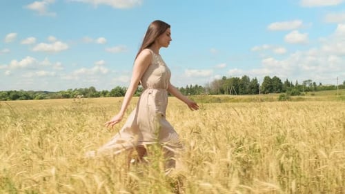 Attractive Young Woman in White Dress Runs Across Yellow Field Against Blue Sky with Clouds
