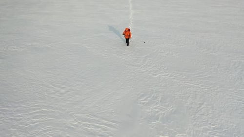 Aerial a Man with a Backpack and Sticks Walks Through the Snowy Desert Leaving a Chain of Footprints