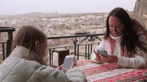 Mother and Daughter Sit at a Cafe Table on the Outdoor Terrace and Watch Their Mobile Phones