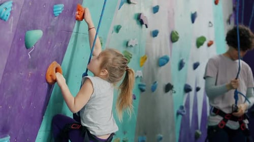 Small Kid Climbing Artificial Wall in Sports Center, Woman Using Safety Equipment in Background