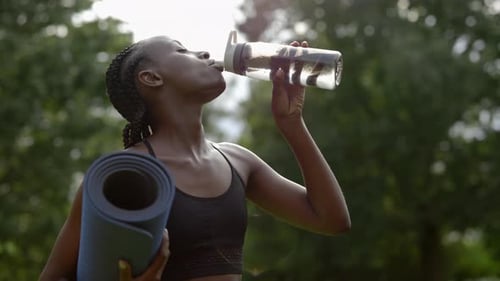 Woman Holding Yoga Mat and Drinking Water at Park